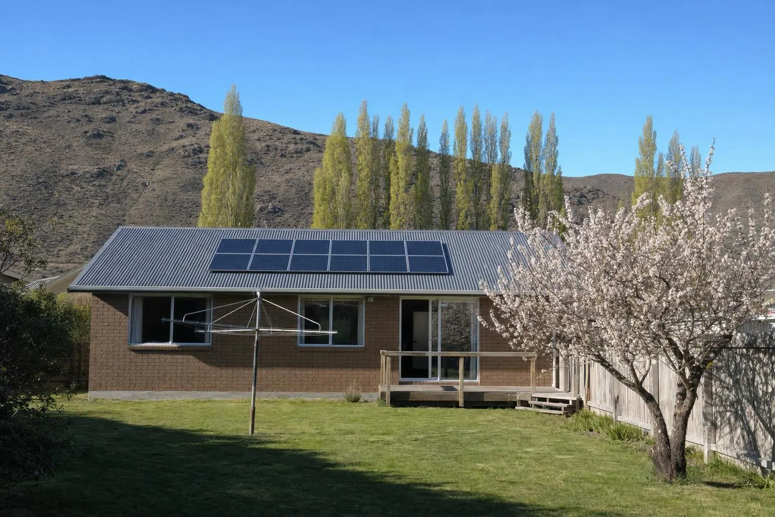 A single-storey brick house with a charcoal iron roof and twelve solar panels, a fruit tree in blossom in the back garden, and poplars and bare rocky hills behind