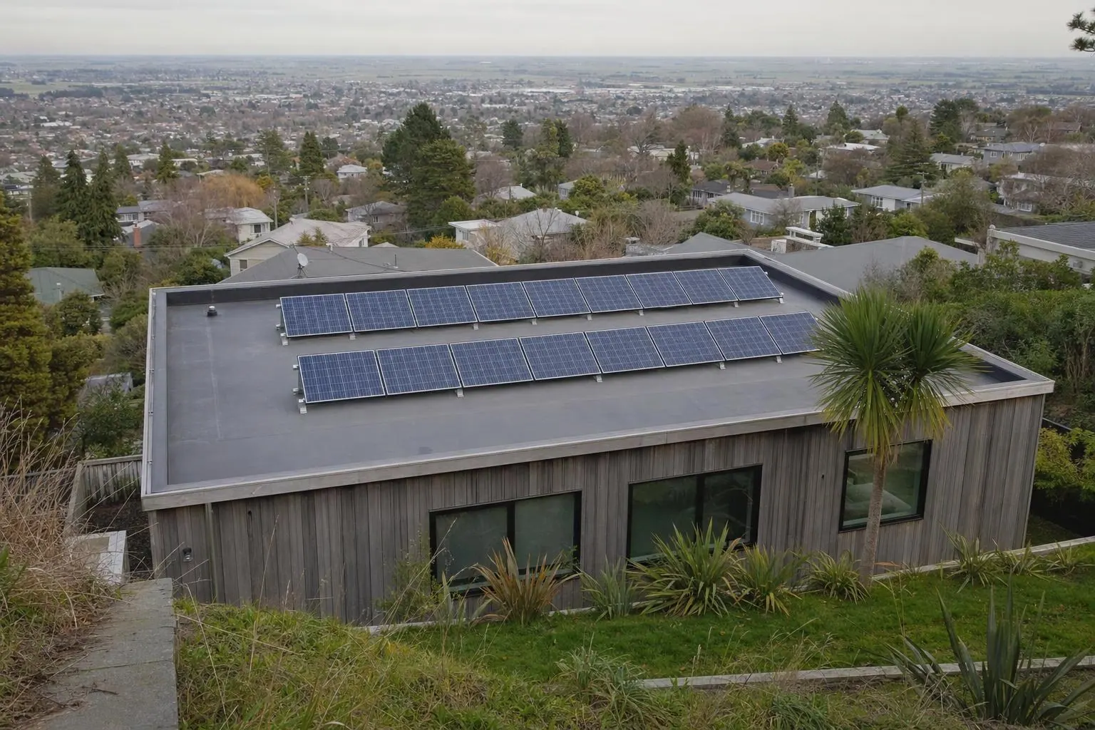 A modern flat-roofed house seen from the street above, with two rows of solar panels tilted up on low frames on its dark membrane roof and suburban rooftops beyond