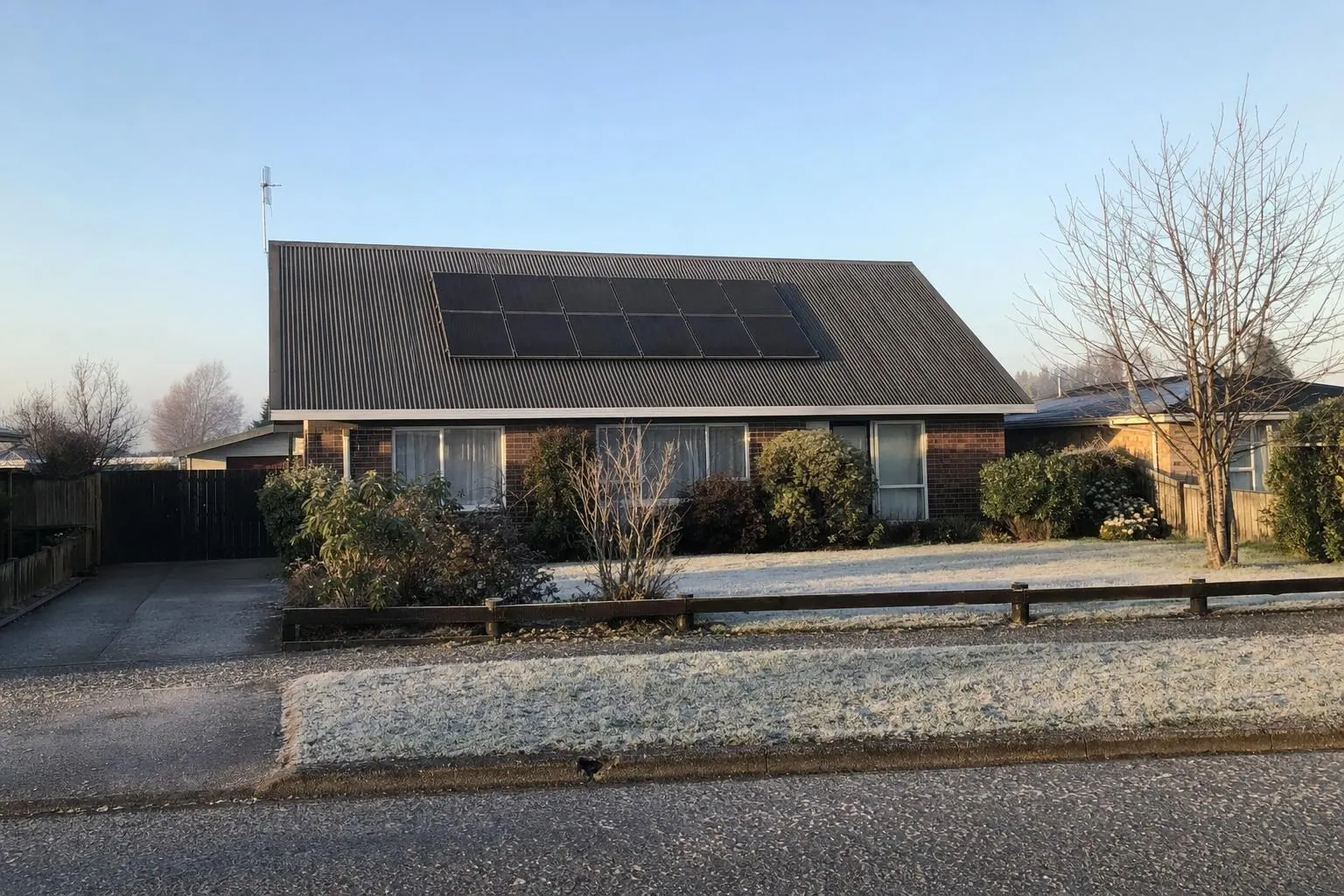 A single-storey brick house with solar panels on its metal roof on a frosty winter morning, with frost on the front lawn and a bare tree beside it