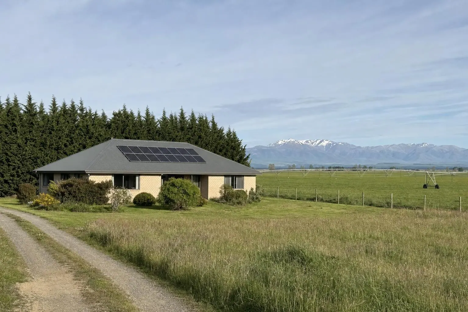 A brick farmhouse with solar panels on its metal roof, a shelter belt behind and a centre-pivot irrigator across the paddock