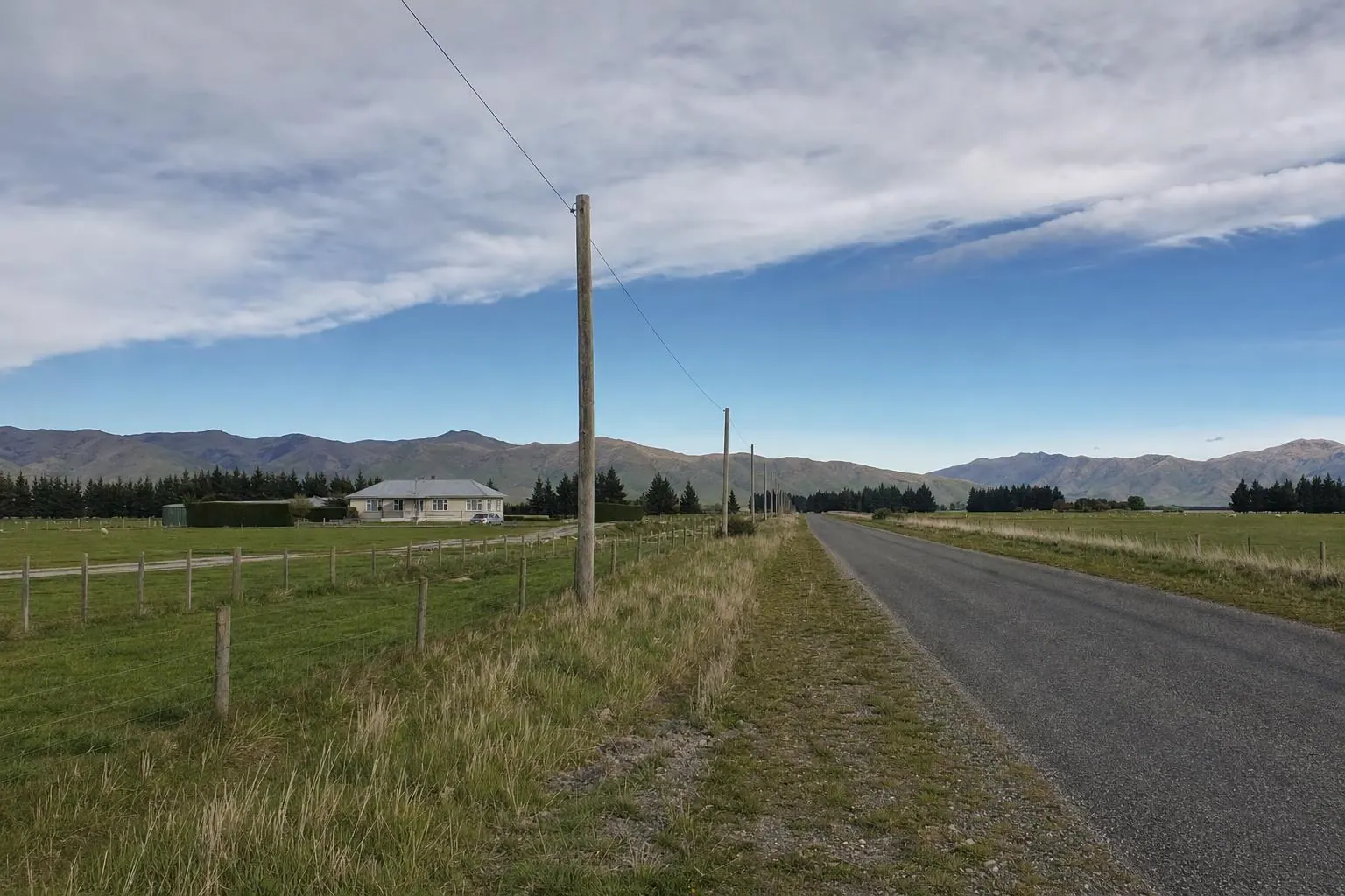 A single-wire power line on wooden poles beside a straight country road, with a farmhouse set back across sheep paddocks and hills in the distance
