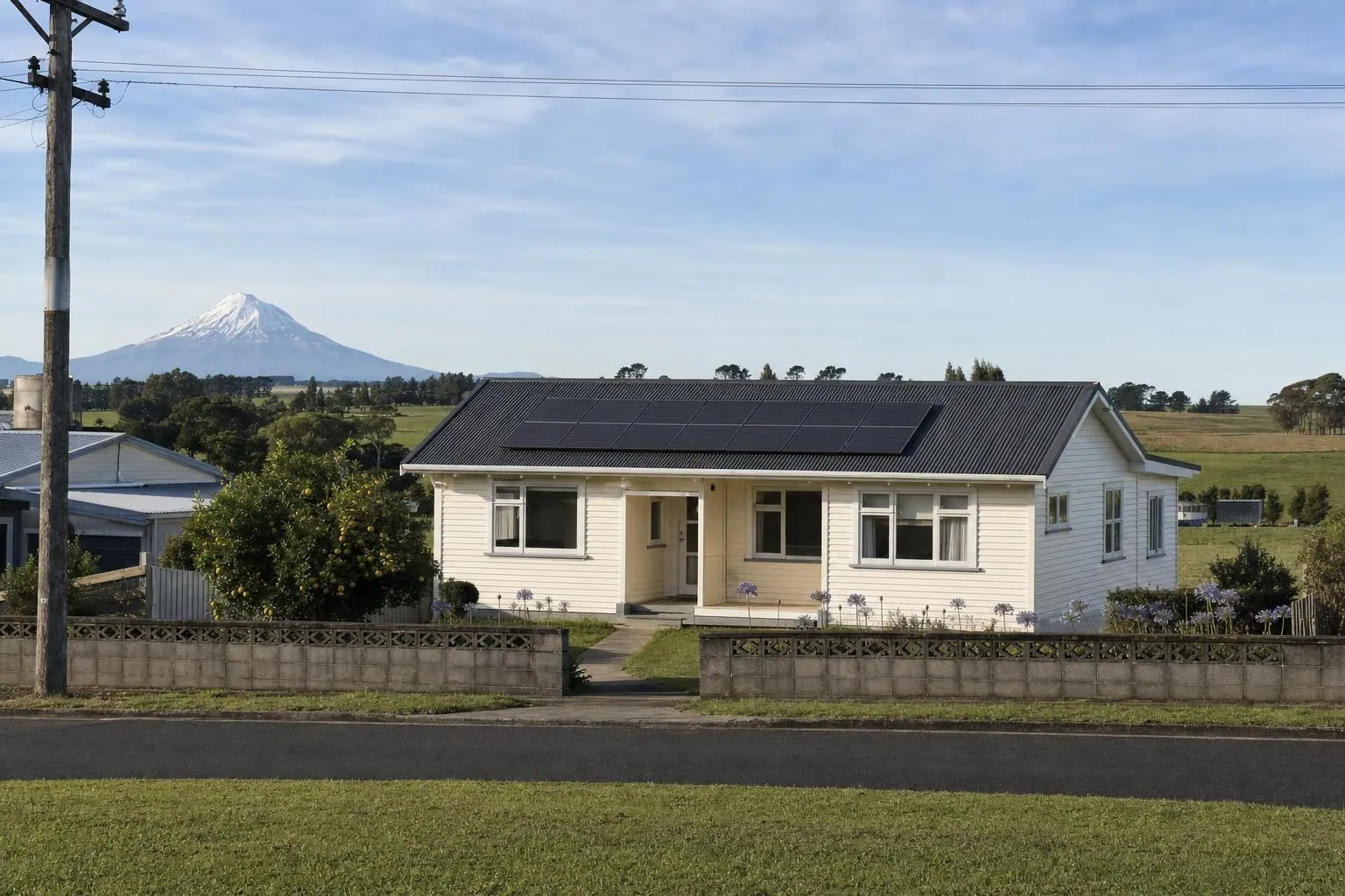 A cream weatherboard bungalow with fourteen solar panels on its charcoal iron roof, green paddocks behind and a snow-capped volcanic cone on the horizon