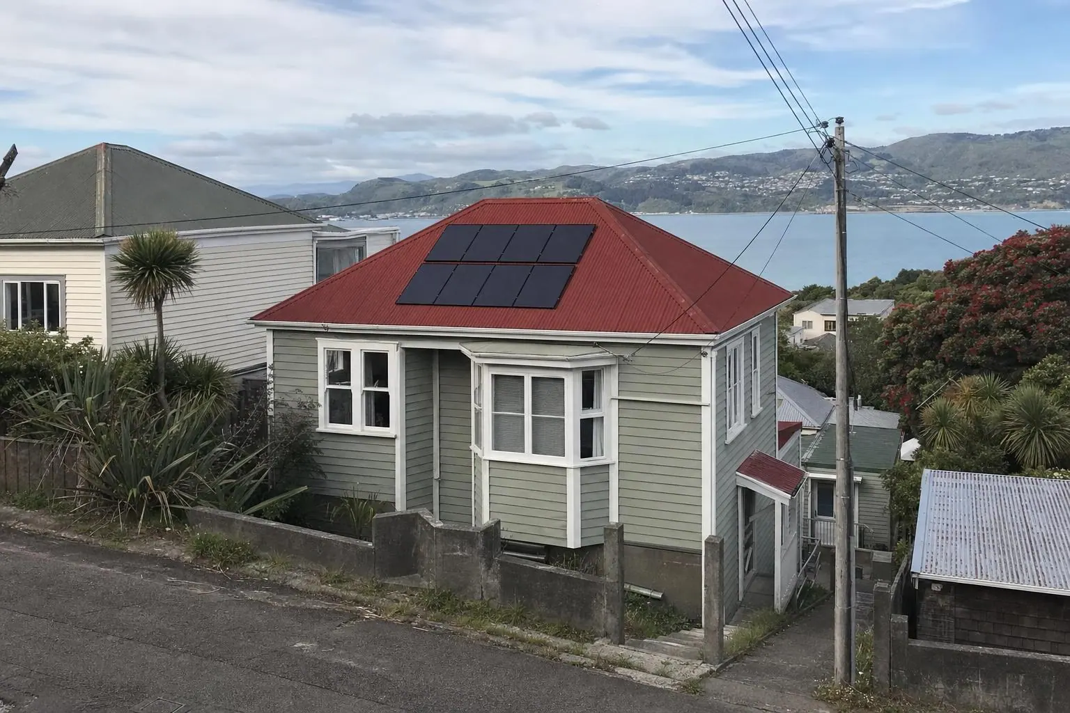 A grey-green weatherboard villa on a steep Wellington street with eight solar panels on its red iron roof, neighbouring houses stepping down the hill and the harbour in the distance